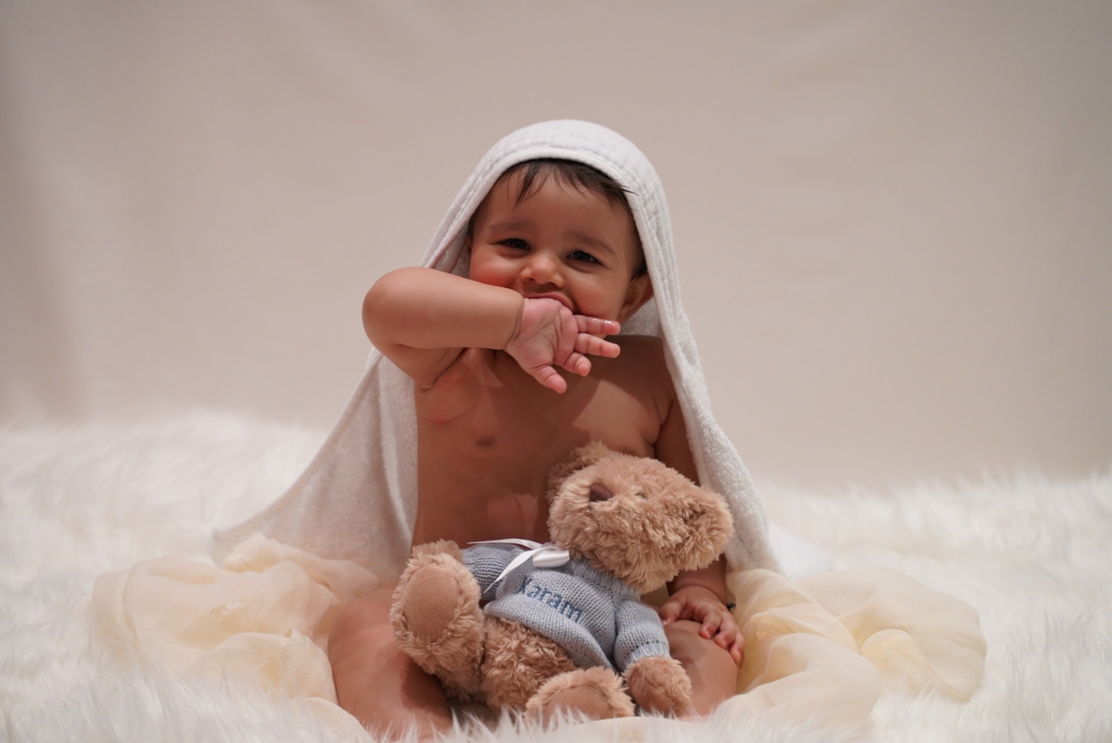 Baby smiling beneath a soft wrap beside a teddy bear